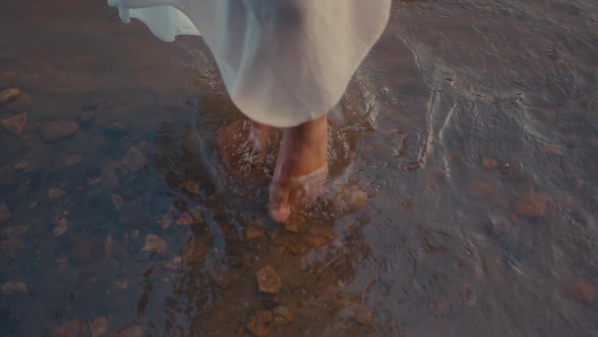 A woman in a dress swirls her foot around in shallow water