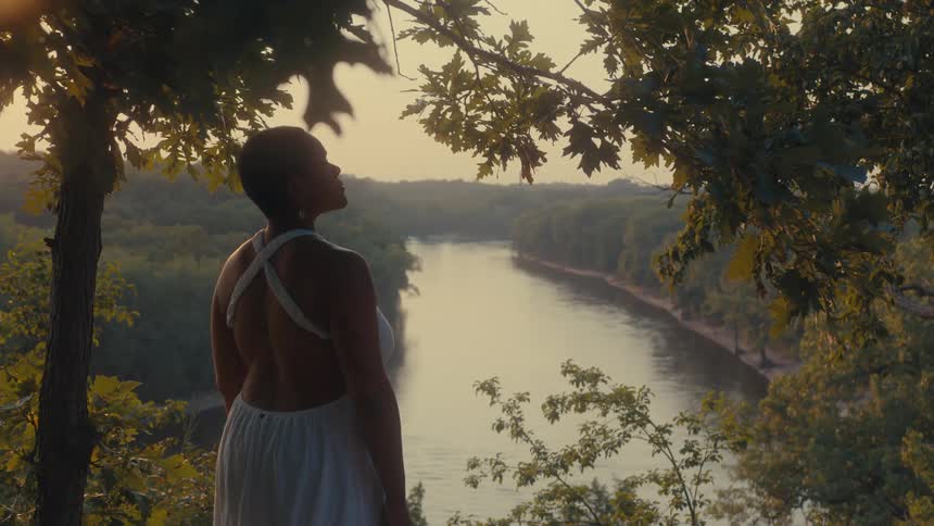 Woman standing at a high point overlooking a river passing through forest looks upward before walking away