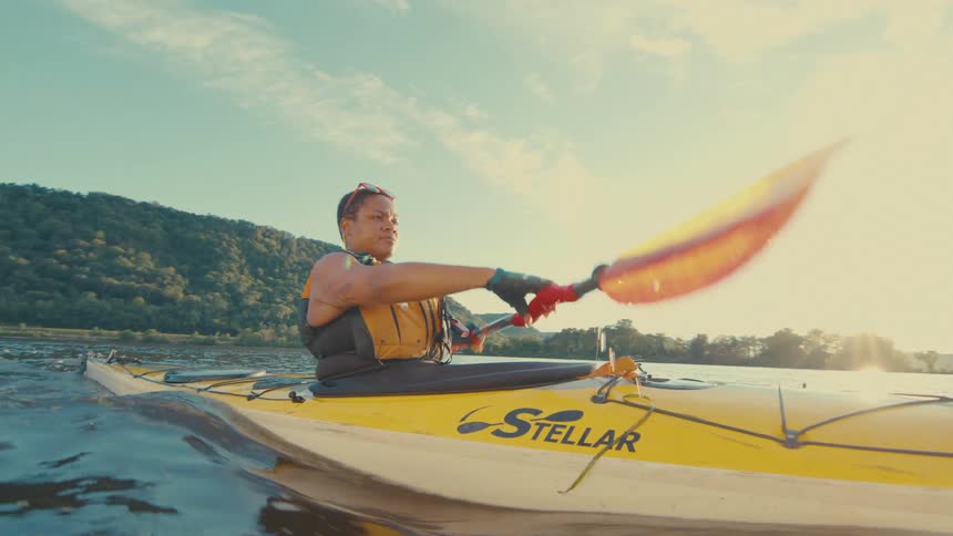 Person paddles a kayak through the water with a forest covered hill in the background