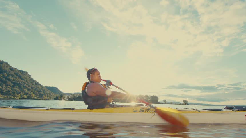 A woman is kayaking on a calm lake with a mountainous landscape in the background during a sunny day