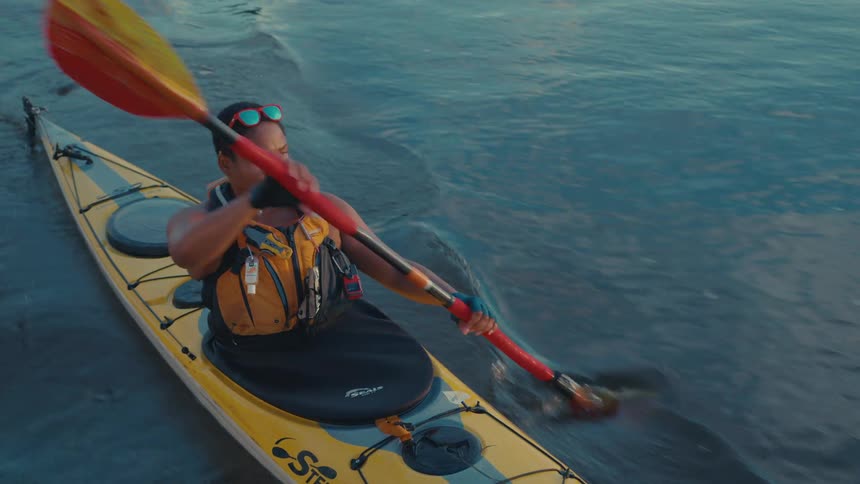 A man using a rudder to kayak in calm water and he is wearing a life jacket