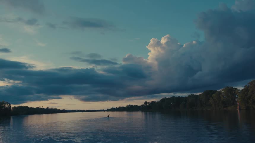 Serene landscape of a river at dusk with thick clouds reflecting the soft light of the sky and a person rowing alone