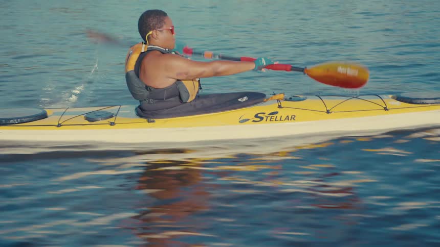 A person wearing a life vest and red sunglasses is kayaking on a lake with trees in the background paddling the yellow and white boat with a red and black oar