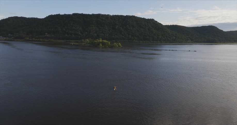 A lone kayaker paddles on a calm expansive river with a densely forested hill under a clear sky
