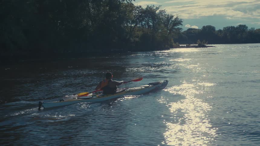 A person is kayaking on a river with a silhouetted tree line and a bright sun reflecting water surface