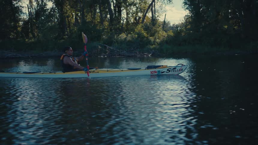 A person wearing a life jacket paddles a yellow and white kayak down a river with trees lining the bank