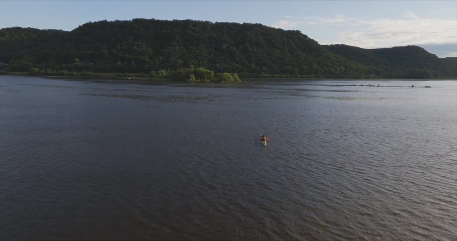 An aerial view of a person kayaking in a bay of water with calm water and a hill of trees in front