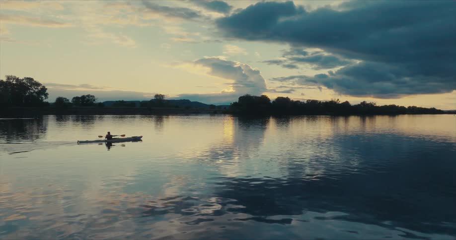 Person paddles a kayak across reflective water towards a red tinted horizon under a cloudy sky