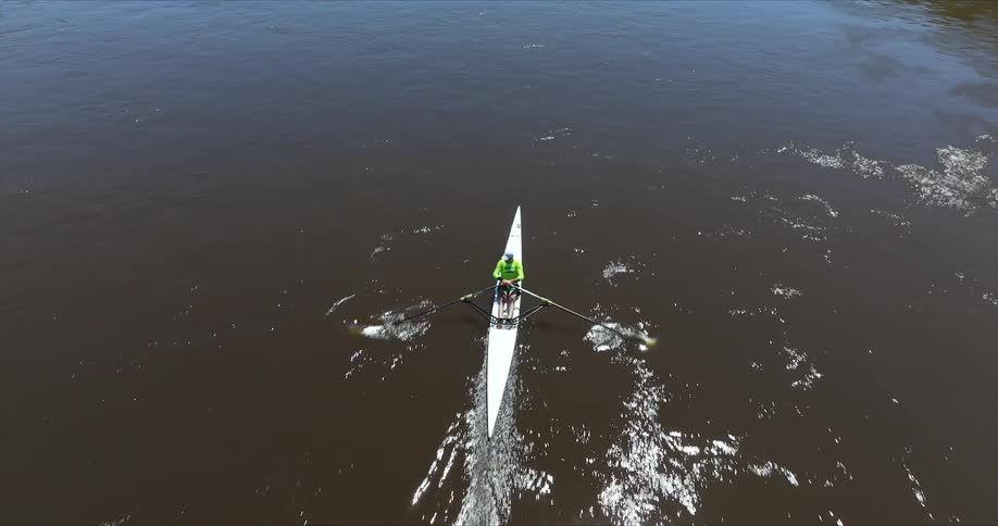 A single sculler rows a white boat down a dark river creating ripples as they move