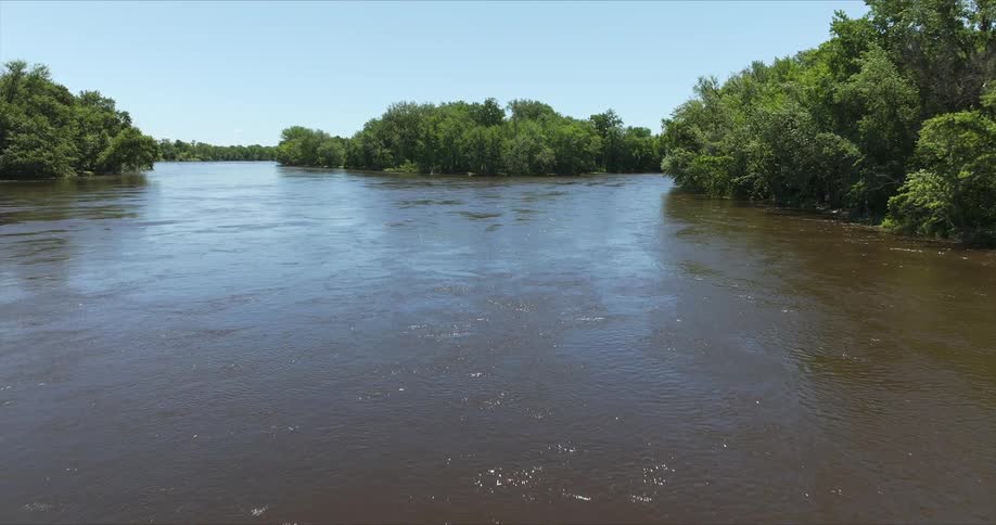 A wide brown river flows through a lush green landscape under a bright blue sky