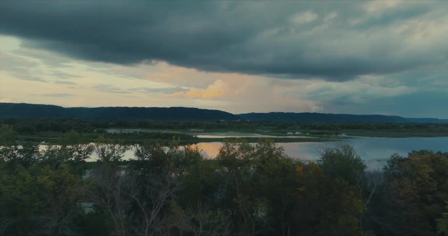 A serene landscape featuring a calm body of water reflecting the cloudy sky with distant hills and lush greenery in the foreground