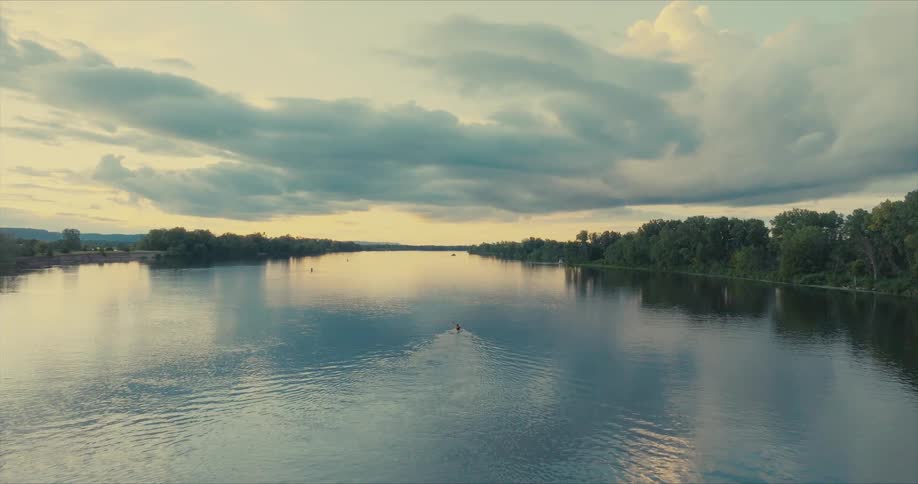 Aerial view of a wide calm river with a dramatic sky of thick clouds and golden reflections at dusk where a lone rower navigates in the center