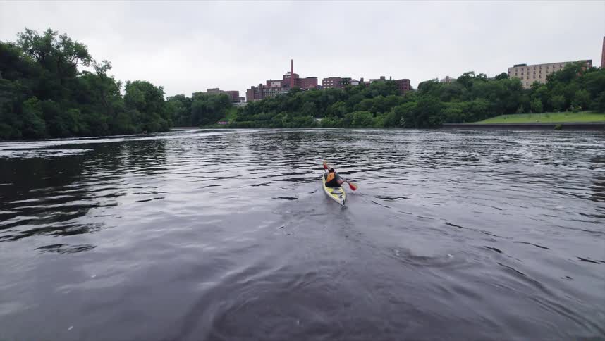 Person paddling a canoe on a river surrounded by vegetation and buildings in the background