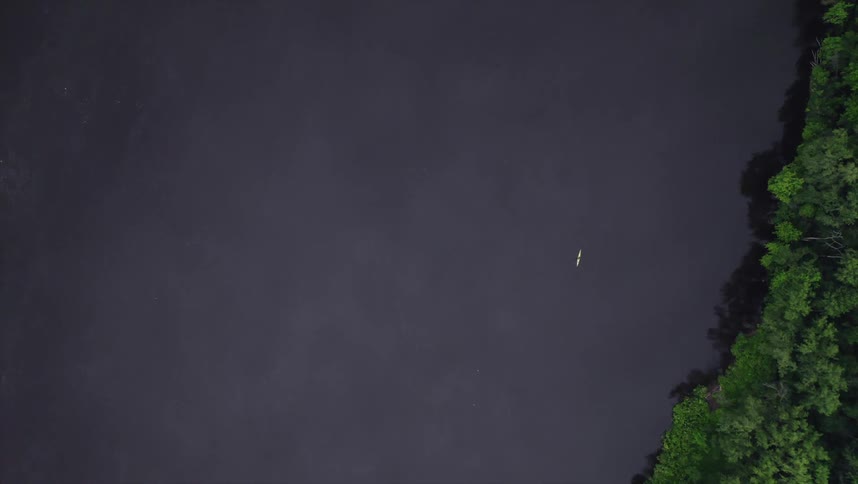 An aerial view shows a lone kayaker paddling across a vast dark lake surrounded by a dense green forest