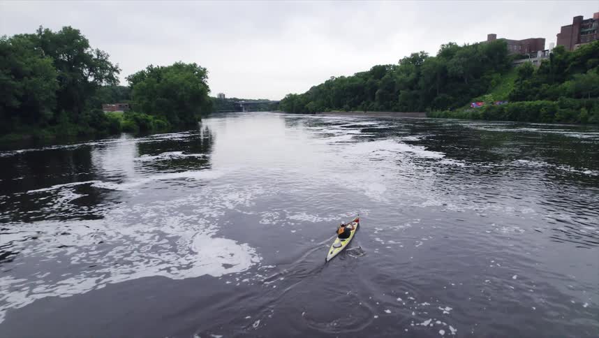 Person paddles a kayak along a river past multiple buildings during the daytime