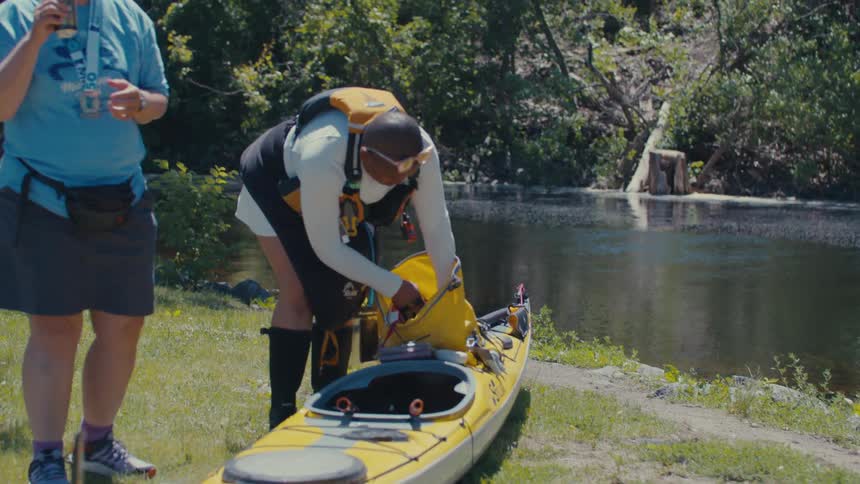A man is taking something out of a yellow bag that is on top of a kayak and is talking to a woman who is next to him