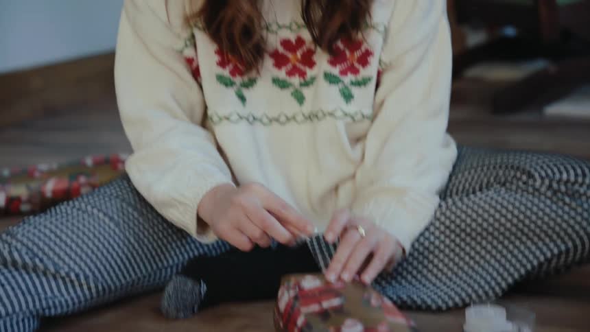 A woman sits on a wooden floor and wraps a small christmas present