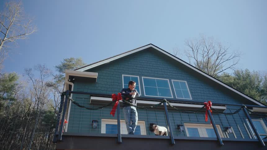 A person is standing with their dog on the balcony of a twostory house decorated with garlands and red bows with a clear blue sky and trees in the background