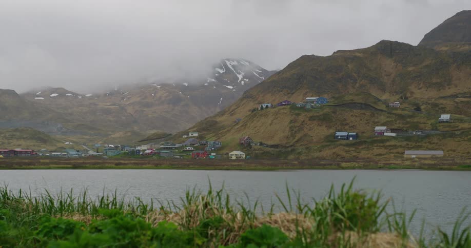 A small remote village with colorful houses nestled in a mountainous landscape with patches of snow situated by a calm lake under a cloudy sky