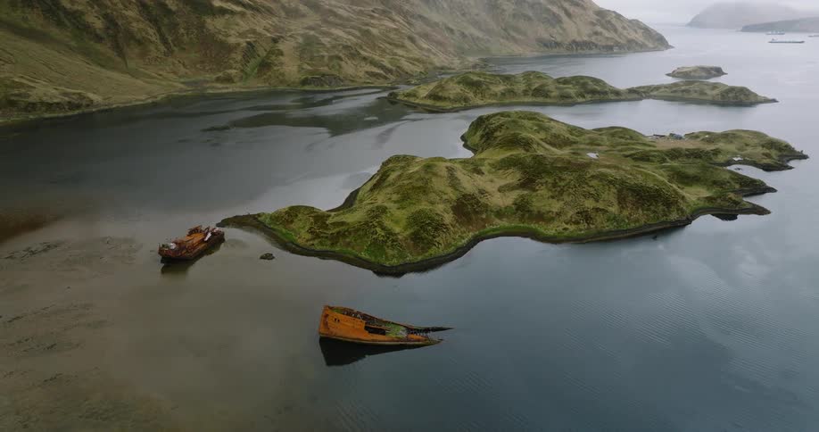 Two shipwrecks lie partially submerged in a calm bay surrounded by lush green hills