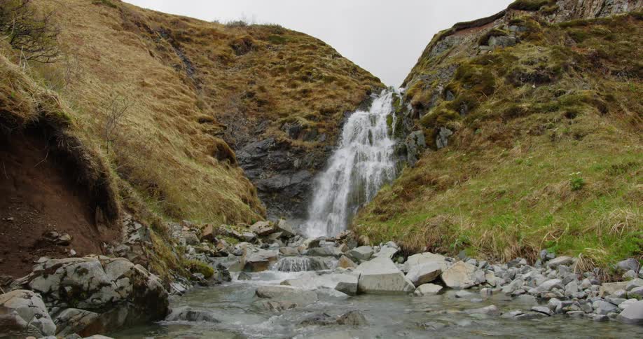 A cascading waterfall plunges down a rocky cliff face surrounded by lush green hillsides and a stream winding through the valley