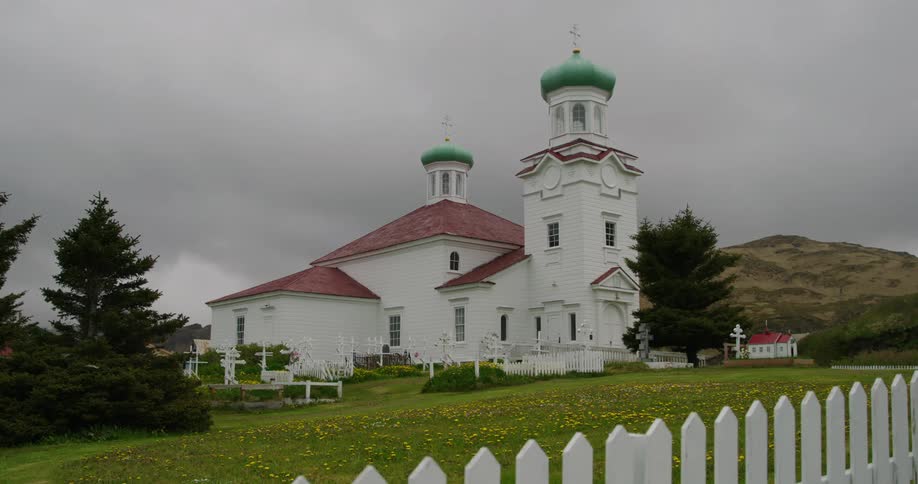 A white church with a green dome stands on a grassy hill surrounded by a picket fence and a small cemetery against a backdrop of a cloudy sky