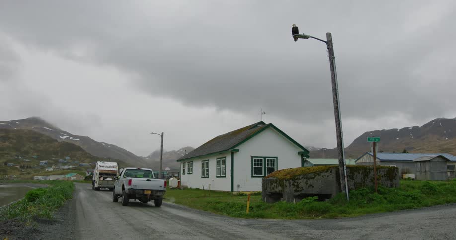 A white garbage truck and a white pickup truck are driving on a gravel road past a small white house
