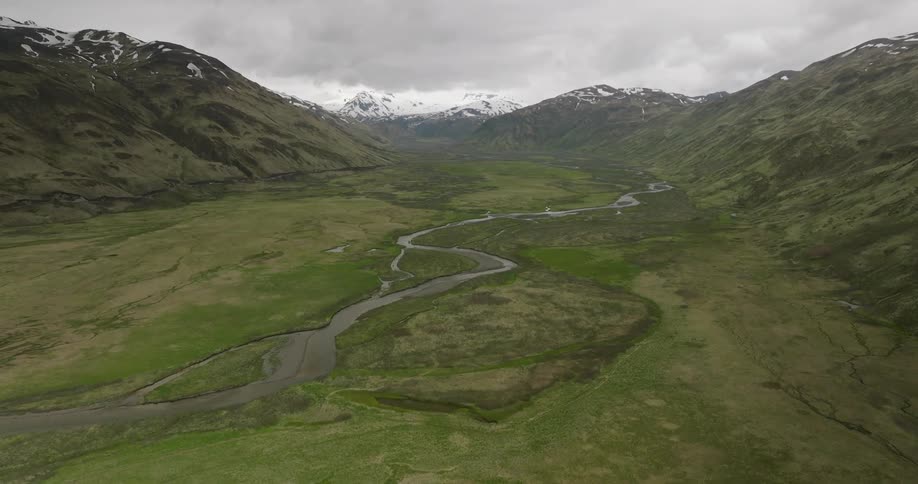 A winding river flows through a lush green valley flanked by snowcapped mountains under a cloudy sky
