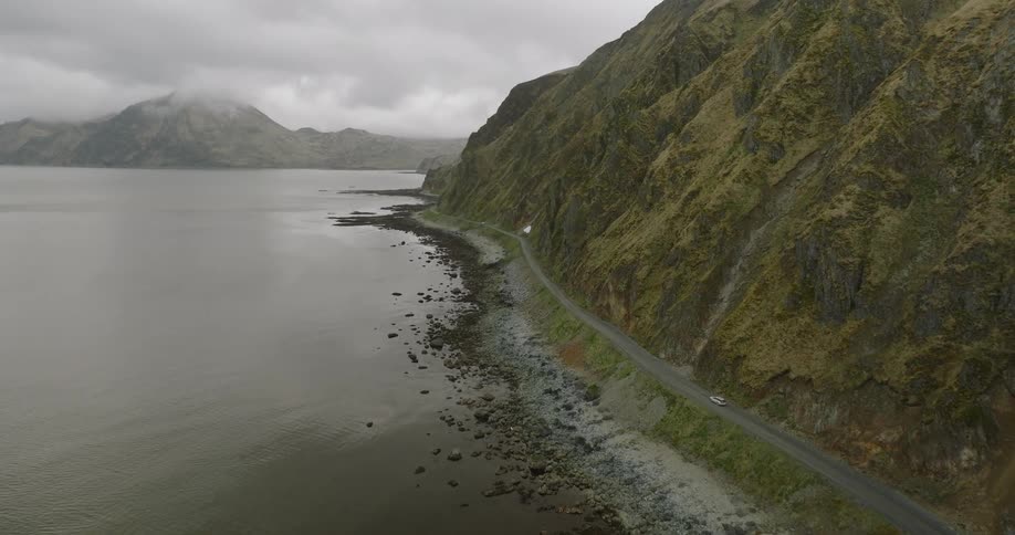 A car driving along a road nestled between a rocky coast and a mossy mountain range all under a cloudy sky