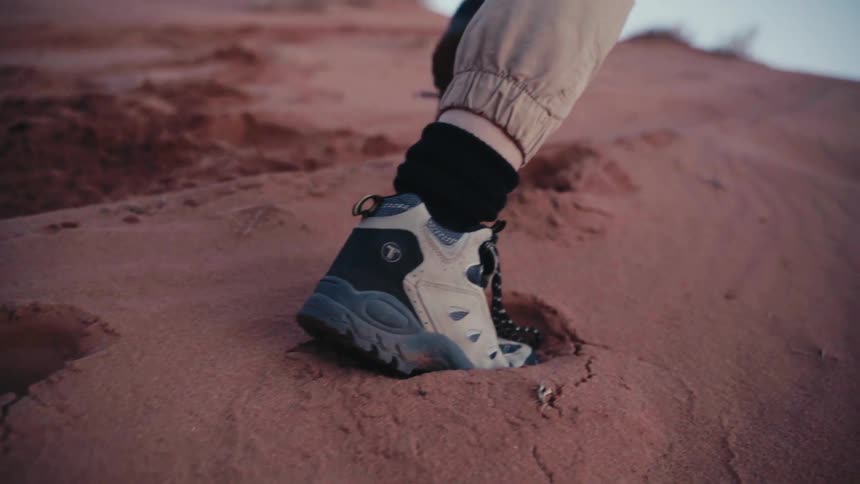 A person wearing hiking boots and beige pants is climbing a sandy hill leaving footprints in the reddish sand