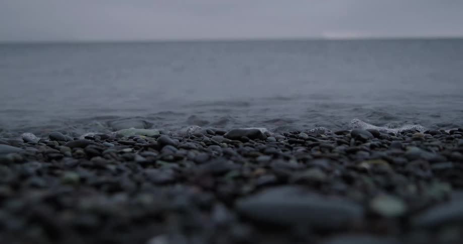 The rough dark pebbles of the beach are smoothed by the relentless crashing of the waves the sea churning and frothing under a stormy grey sky sending a spray of icy water across the shore