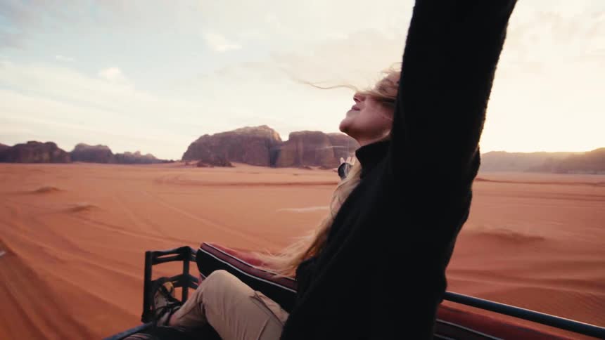 Blonde woman in the back seat of a car spreading her arms as they move through a desert area with mountains in the background