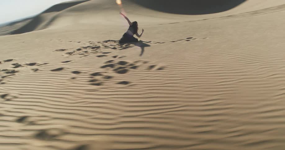 A person sits on a vast undulating sand dune leaving a trail of footprints behind while dancing