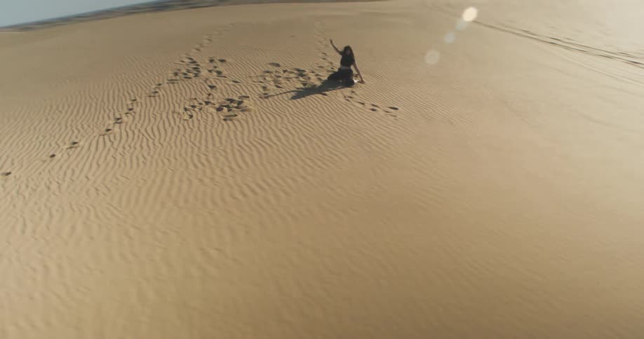A person sits on a vast undulating sand dune leaving a trail of footprints behind and tossing sand into the air