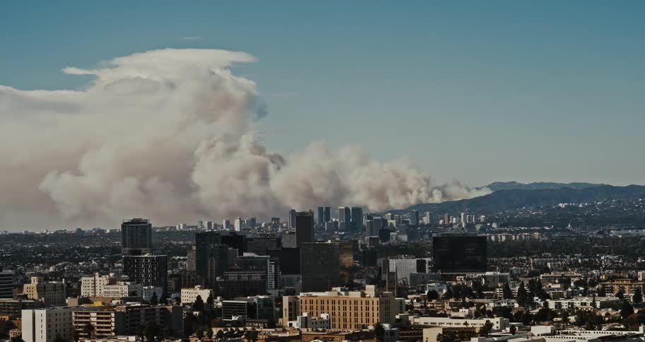 Los Angeles cityscape with skyscrapers in the foreground stretches towards the Santa Monica mountains where a thick column of smoke rises into the sky