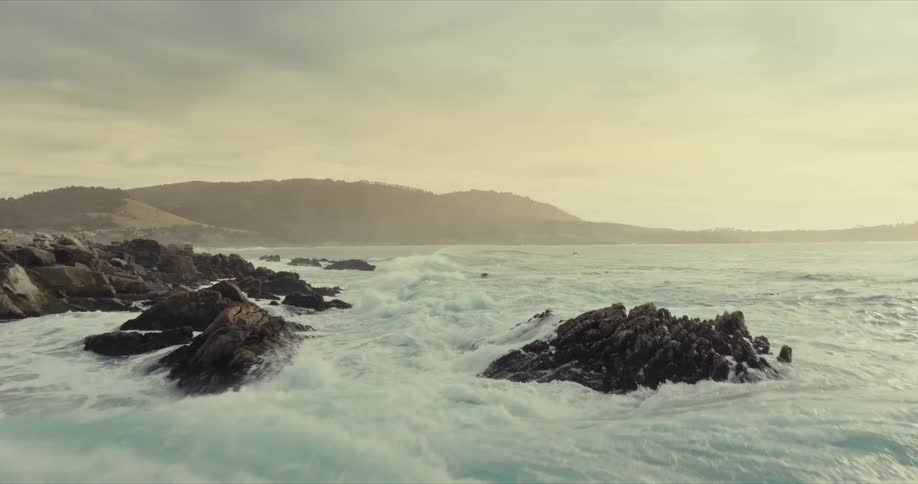 A rocky coastline with crashing waves and a hazy sunset over the horizon