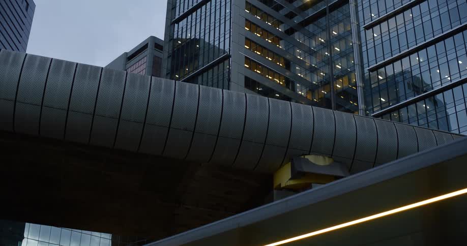 A busy city street at dusk where towering skyscrapers with reflective glass facades dominate the skyline their windows aglow with warm light and a pedestrian walkway stretches across the street connecting buildings on either side
