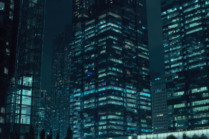 A cluster of skyscrapers at night with their glass facades reflecting the city lights and the windows turning on in a flickering pattern