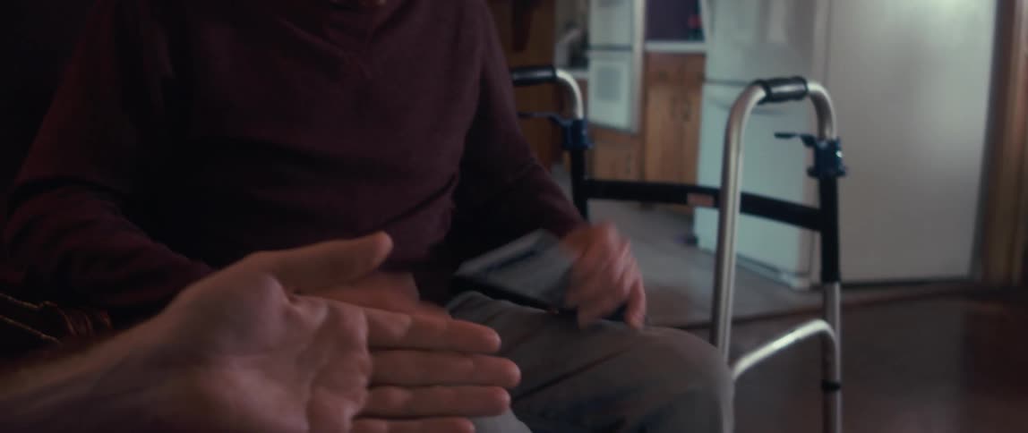 A male nurse in a blue shirt shows brain scan images to an elderly man sitting in a chair with a walker nearby in a cozy woodpaneled room
