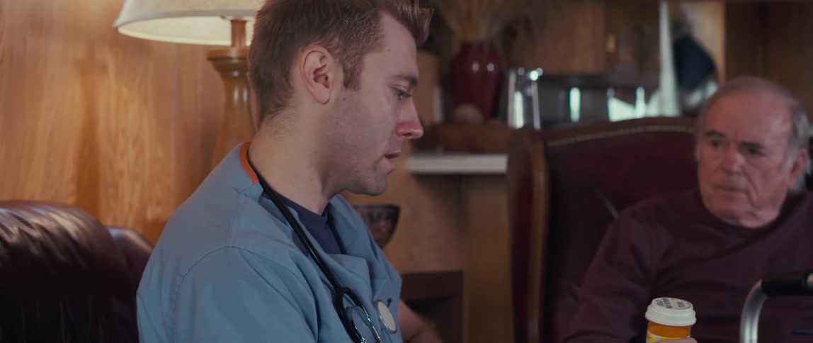 A male nurse in a blue shirt shows prescription pill bottles to an elderly man sitting in a chair with a walker nearby in a cozy woodpaneled room