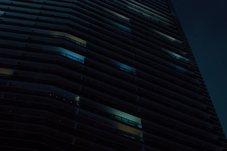 A towering contemporary building adorned with a multitude of windows some illuminated in various hues creating a dynamic pattern as the lights flicker before turning off against the backdrop of a dark nighttime sky