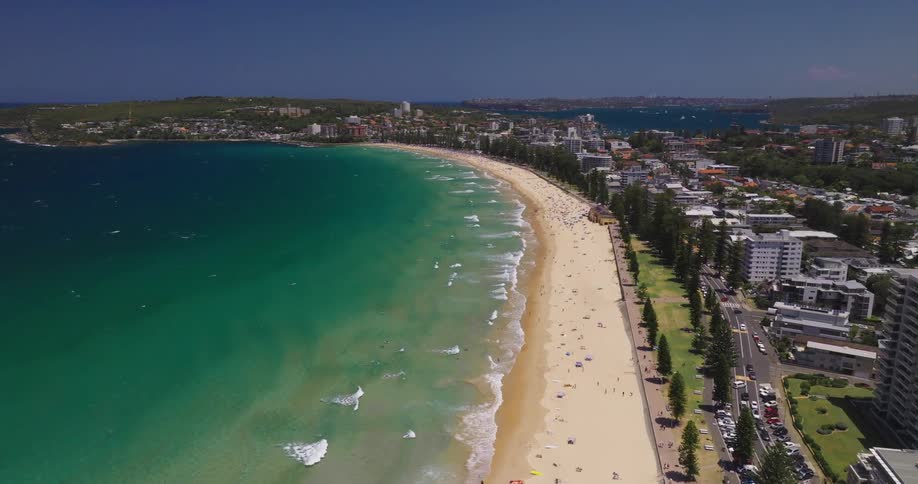 An aerial view of a long sandy beach lined with buildings and dotted with sunbathers with the turquoise ocean stretching out beyond