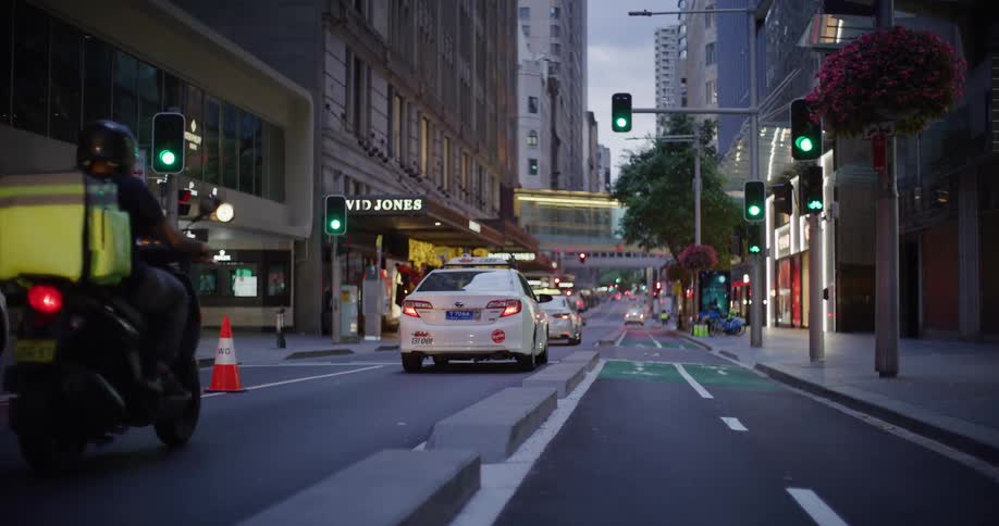 White cabs and delivery motorcycles navigate light traffic on a city street at dusk