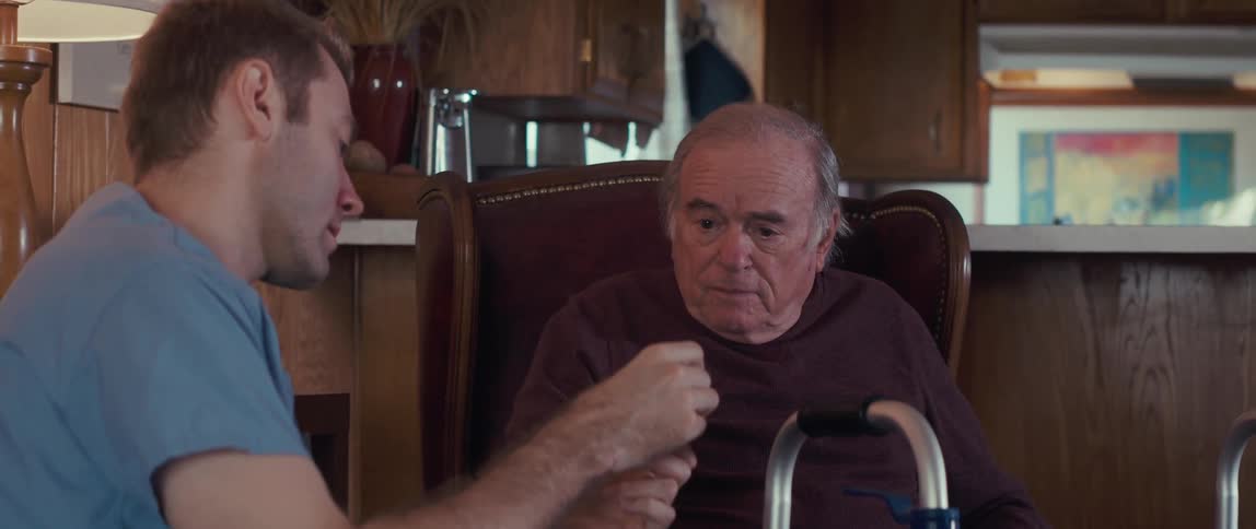 Nurse taking a medicine and showing it to an elderly man who is sitting next to him in an armchair
