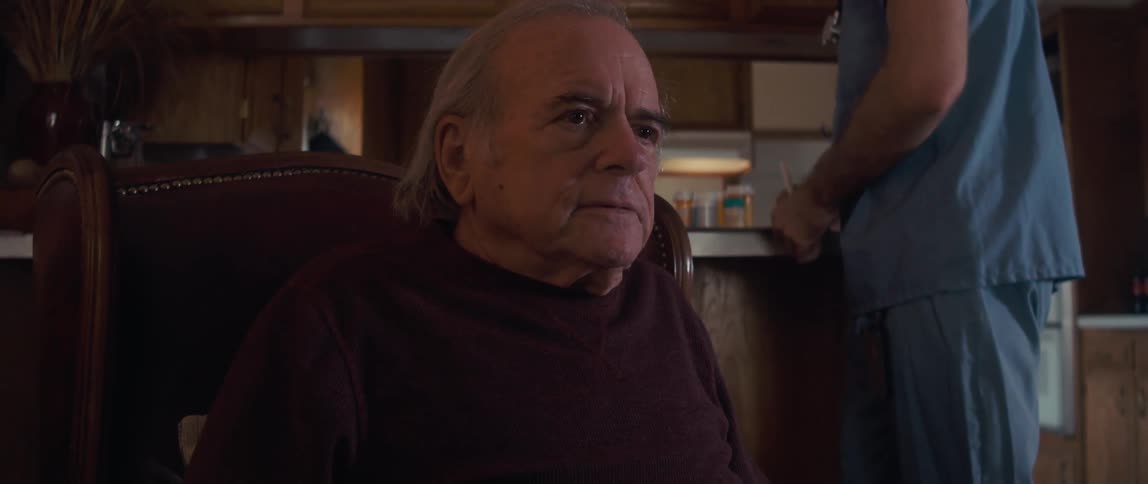 Elderly man sitting in an armchair with a nurse in the background
