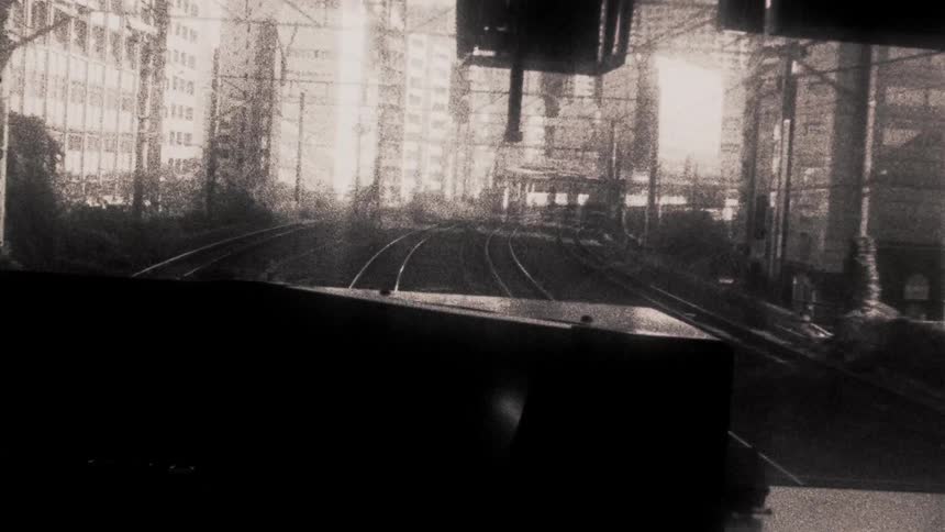 A view from the front of a train moving through an urban area with multiple tracks surrounded by tall buildings and overhead electrical wires