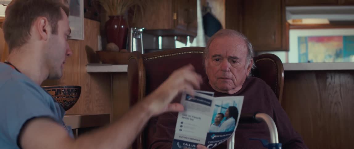 A male nurse in a blue shirt shows a brochure to an elderly man sitting in a chair with a walker nearby in a cozy woodpaneled room