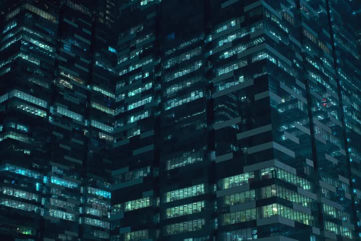 A cluster of densely packed skyscrapers bathed in a cool blue light casts a mesmerizing glow against the dark night sky as their glass facades reflect the citys illumination