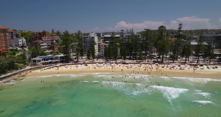 A busy beach with people swimming and sunbathing surrounded by trees and buildings