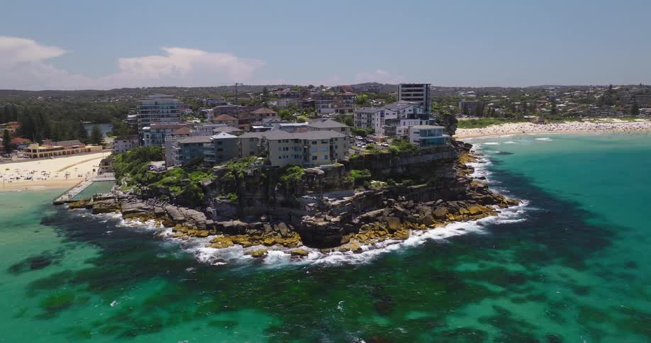 A coastal scene with a rocky outcrop featuring several buildings surrounded by turquoise waters and sandy beaches